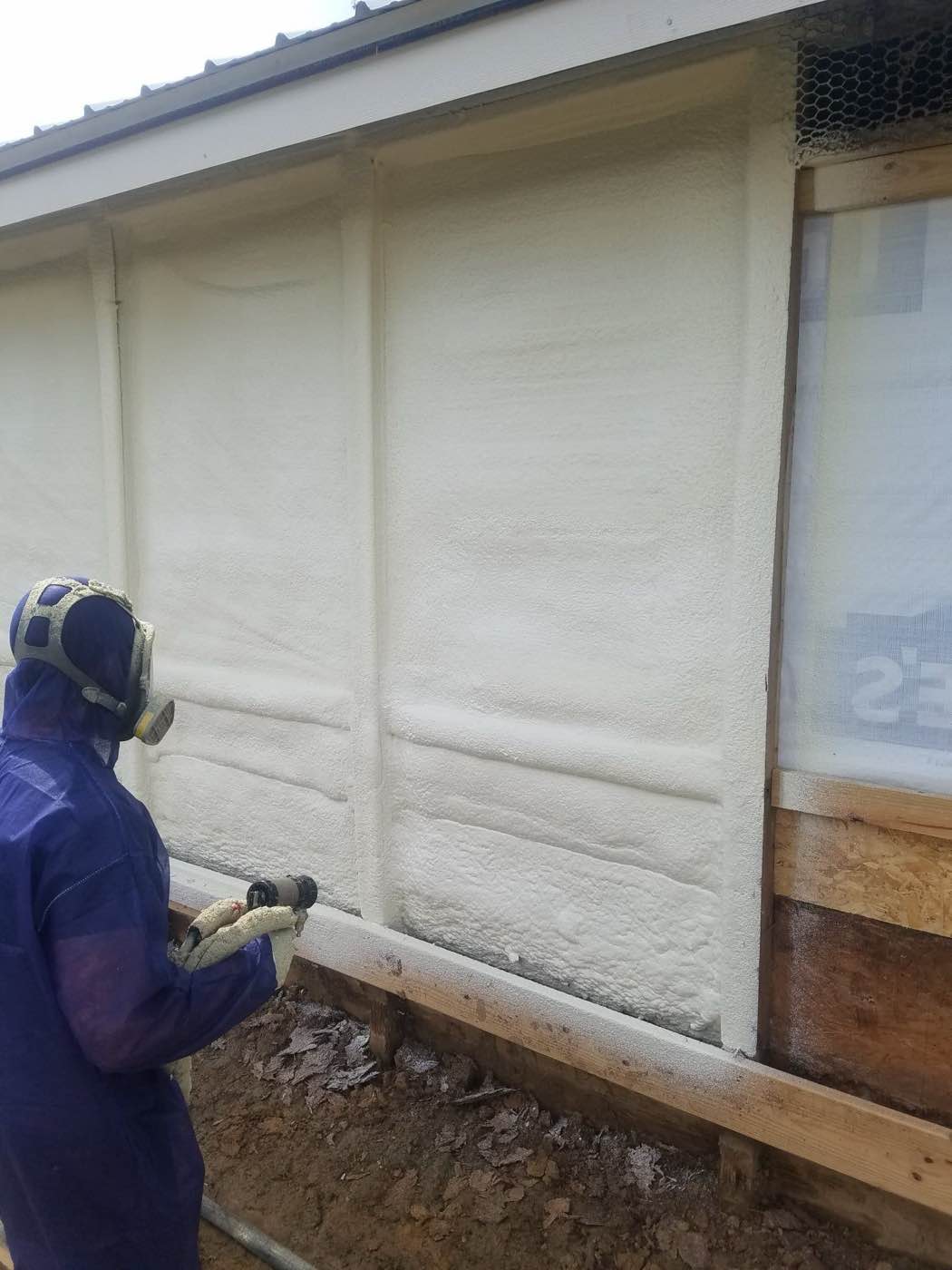 Insulation technician in protective gear spraying foam along an interior wall