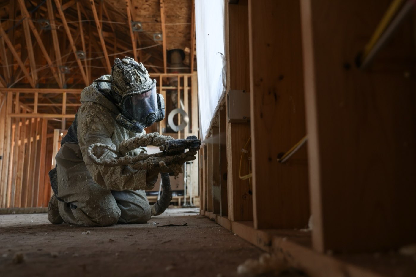 A spray foam insulation technician in full protective gear spraying closed cell foam into wall framing